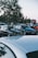 A group of parked cars is seen in an outdoor setting typical of a car meet. People are gathered around, some standing near the cars and conversing. The vehicles are older models, with visible details like decals and custom features. Trees and a clear sky form the backdrop, creating a relaxed and social atmosphere.