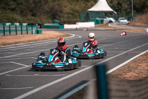 Two individuals are racing go-karts on an outdoor track. The track is paved and features several curves, with green barriers along the sides. The racers wear helmets and protective gear, and the vehicles display numbers and vibrant colors. The background includes some greenery and a white tent structure.
