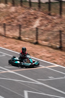 A person wearing a helmet is driving a go-kart on an outdoor track. The go-kart is black and teal, marked with the number 23. The track is outlined with white lines, and the surroundings include a dry, brown landscape with a fence.