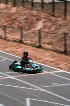 A person wearing a helmet is driving a go-kart on an outdoor track. The go-kart is black and teal, marked with the number 23. The track is outlined with white lines, and the surroundings include a dry, brown landscape with a fence.