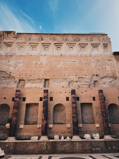 A historic brick structure featuring a series of tall, cylindrical columns adorned with variegated patterns. The wall above displays a sequence of recessed, square designs and exhibits a weathered texture. The foreground includes a patterned stone floor, remnants of sculptural pieces, and additional architectural elements.