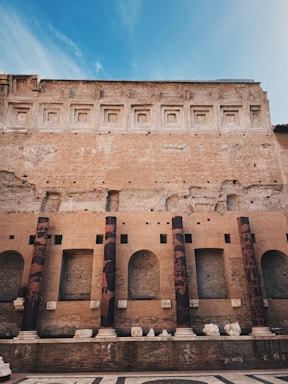 A historic brick structure featuring a series of tall, cylindrical columns adorned with variegated patterns. The wall above displays a sequence of recessed, square designs and exhibits a weathered texture. The foreground includes a patterned stone floor, remnants of sculptural pieces, and additional architectural elements.