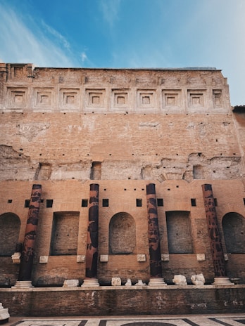 A historic brick structure featuring a series of tall, cylindrical columns adorned with variegated patterns. The wall above displays a sequence of recessed, square designs and exhibits a weathered texture. The foreground includes a patterned stone floor, remnants of sculptural pieces, and additional architectural elements.