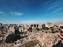 Ancient ruins spread across a vast area, featuring classical Roman architecture with columns and arches. A blue sky stretches overhead, and a few people are visible in the foreground, appearing to take pictures or look at the scenery.