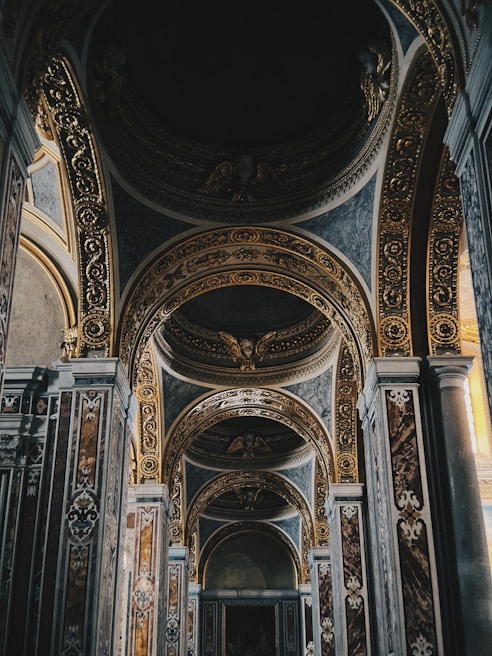 An intricately decorated interior with ornate arches and golden embellishments. The design features detailed carvings and symmetrical patterns, enhancing the grandeur of the space. The lighting appears soft, creating a serene and majestic atmosphere.