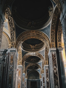 An intricately decorated interior with ornate arches and golden embellishments. The design features detailed carvings and symmetrical patterns, enhancing the grandeur of the space. The lighting appears soft, creating a serene and majestic atmosphere.