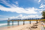 A sunny beach scene featuring a sign stretching across the sand with letters spelling out 'LABUHAN JUKUNG KRUI'. The ocean waves gently lap at the shore under a vibrant blue sky scattered with clouds. A couple of people walk along the sandy beach, and palm trees can be seen in the background.