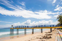 A sunny beach scene featuring a sign stretching across the sand with letters spelling out 'LABUHAN JUKUNG KRUI'. The ocean waves gently lap at the shore under a vibrant blue sky scattered with clouds. A couple of people walk along the sandy beach, and palm trees can be seen in the background.