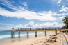 A sunny beach scene featuring a sign stretching across the sand with letters spelling out 'LABUHAN JUKUNG KRUI'. The ocean waves gently lap at the shore under a vibrant blue sky scattered with clouds. A couple of people walk along the sandy beach, and palm trees can be seen in the background.