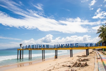 A sunny beach scene featuring a sign stretching across the sand with letters spelling out 'LABUHAN JUKUNG KRUI'. The ocean waves gently lap at the shore under a vibrant blue sky scattered with clouds. A couple of people walk along the sandy beach, and palm trees can be seen in the background.