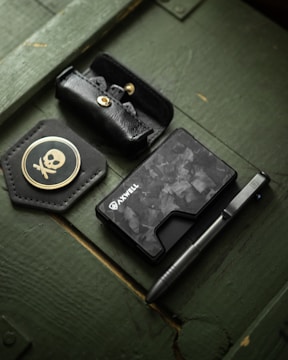 A flat lay showcasing a black leather pouch, a pen, a wallet with a camouflage pattern labeled 'Axwell,' and a black leather patch with a skull and crossbones emblem, all placed on a green wooden surface.