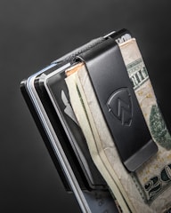 A close-up of a metal money clip holding several bills and cards. The clip is black with a logo, and the bills include U.S. currency. The background is a simple dark gray color, adding a sleek, minimalist look.