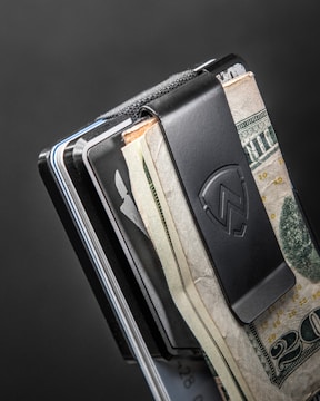 A close-up of a metal money clip holding several bills and cards. The clip is black with a logo, and the bills include U.S. currency. The background is a simple dark gray color, adding a sleek, minimalist look.