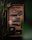 A neatly arranged tray features various black and metallic objects including a tactical pen, a carabiner, a badge with a skull and compass design, a compact flashlight, a stylish dark wallet, a folding knife, and a minimalist phone case. Bamboo plants add a touch of green.