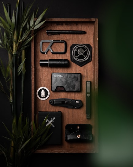 A neatly arranged tray features various black and metallic objects including a tactical pen, a carabiner, a badge with a skull and compass design, a compact flashlight, a stylish dark wallet, a folding knife, and a minimalist phone case. Bamboo plants add a touch of green.