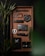 A neatly arranged tray features various black and metallic objects including a tactical pen, a carabiner, a badge with a skull and compass design, a compact flashlight, a stylish dark wallet, a folding knife, and a minimalist phone case. Bamboo plants add a touch of green.