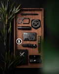 A neatly arranged tray features various black and metallic objects including a tactical pen, a carabiner, a badge with a skull and compass design, a compact flashlight, a stylish dark wallet, a folding knife, and a minimalist phone case. Bamboo plants add a touch of green.