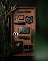 A neatly arranged tray features various black and metallic objects including a tactical pen, a carabiner, a badge with a skull and compass design, a compact flashlight, a stylish dark wallet, a folding knife, and a minimalist phone case. Bamboo plants add a touch of green.
