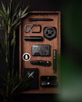 A neatly arranged tray features various black and metallic objects including a tactical pen, a carabiner, a badge with a skull and compass design, a compact flashlight, a stylish dark wallet, a folding knife, and a minimalist phone case. Bamboo plants add a touch of green.