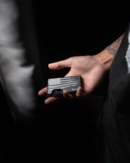 A hand holds a slim black wallet featuring a monochrome American flag design with stars and stripes. The background is dark, focusing attention on the hand and wallet.