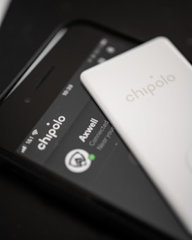 A close-up view of a smartphone displaying the Chipolo app interface, with a white Chipolo tracking device placed on top of the phone. The phone screen shows details like connectivity status, including the name Axwell and a connected status icon.
