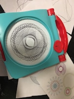 A geometric drawing tool with a turquoise frame and red handles used to create intricate spiral patterns. Several examples of the patterns, in various colors, are seen on white sheets underneath the tool.