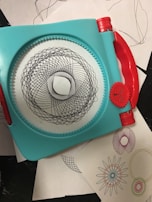 A geometric drawing tool with a turquoise frame and red handles used to create intricate spiral patterns. Several examples of the patterns, in various colors, are seen on white sheets underneath the tool.