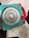 A geometric drawing tool with a turquoise frame and red handles used to create intricate spiral patterns. Several examples of the patterns, in various colors, are seen on white sheets underneath the tool.