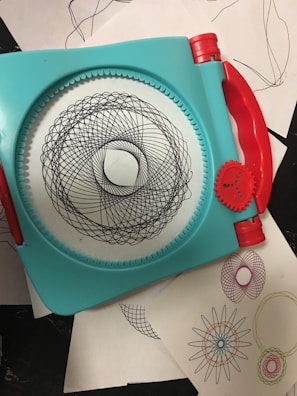 A geometric drawing tool with a turquoise frame and red handles used to create intricate spiral patterns. Several examples of the patterns, in various colors, are seen on white sheets underneath the tool.