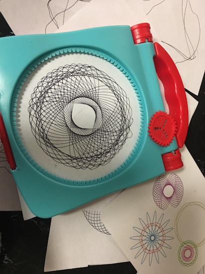 A geometric drawing tool with a turquoise frame and red handles used to create intricate spiral patterns. Several examples of the patterns, in various colors, are seen on white sheets underneath the tool.