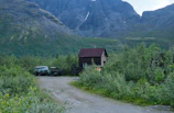 A small wooden cabin is nestled in a lush green forest at the base of a rugged mountain range. An old green vehicle is parked next to a trailer on a dirt path leading to the cabin. The surrounding vegetation appears dense and vibrant, with a backdrop of steep rocky cliffs under a cloudy sky.
