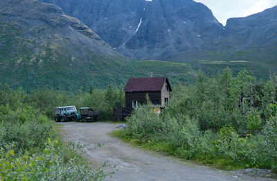 A small wooden cabin is nestled in a lush green forest at the base of a rugged mountain range. An old green vehicle is parked next to a trailer on a dirt path leading to the cabin. The surrounding vegetation appears dense and vibrant, with a backdrop of steep rocky cliffs under a cloudy sky.