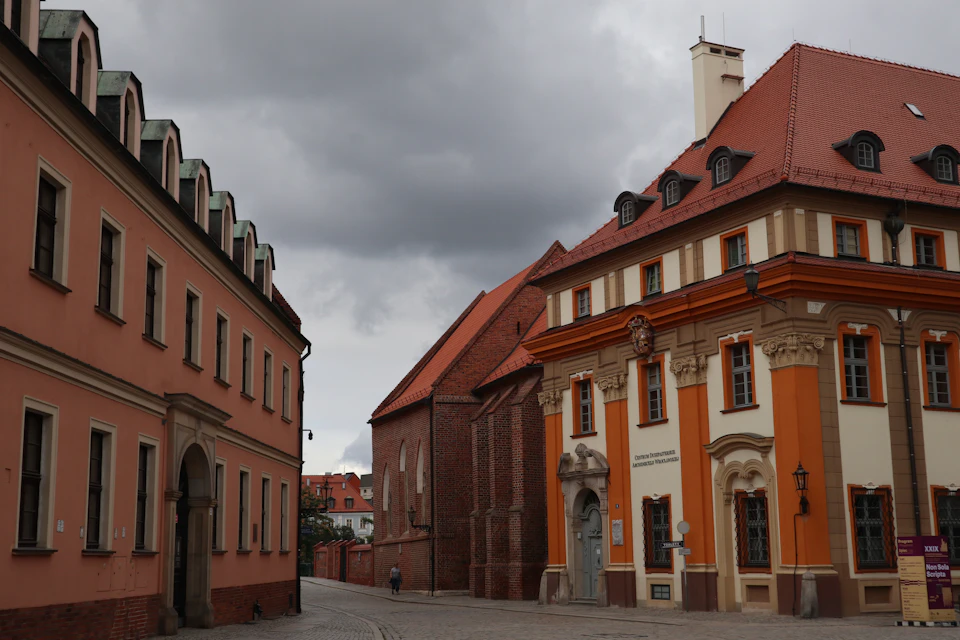 The image depicts a cobblestone street with two buildings on either side, one painted in a warm orange hue and the other in a cool blue shade. The building on the left is adorned with a green roof, while the right-side building features a red roof. A solitary figure can be seen walking along the sidewalk between these structures.