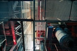 An industrial setting is depicted with metal grates, beams, and various pipes. The structure includes red and blue hues, giving a mechanical appearance. The angle looks upwards, highlighting the intricate grid-like design and machinery components.