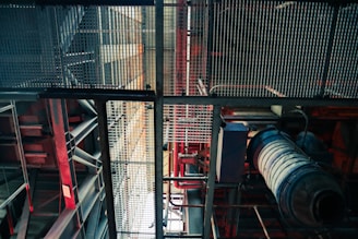 An industrial setting is depicted with metal grates, beams, and various pipes. The structure includes red and blue hues, giving a mechanical appearance. The angle looks upwards, highlighting the intricate grid-like design and machinery components.
