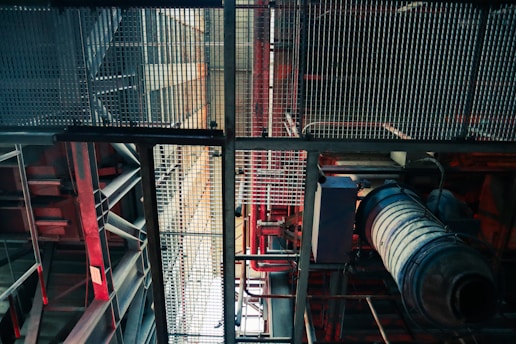 An industrial setting is depicted with metal grates, beams, and various pipes. The structure includes red and blue hues, giving a mechanical appearance. The angle looks upwards, highlighting the intricate grid-like design and machinery components.