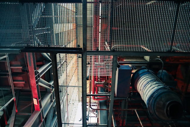 An industrial setting is depicted with metal grates, beams, and various pipes. The structure includes red and blue hues, giving a mechanical appearance. The angle looks upwards, highlighting the intricate grid-like design and machinery components.