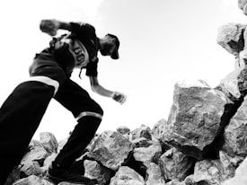 A person in athletic gear appears to be climbing or moving quickly over a large pile of rocks. The image is in black and white, giving it a dramatic and dynamic perspective, with the sky visible in the background. The person's motion is slightly blurred, suggesting movement.
