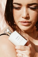 A person with closed eyes is holding a white perfume bottle close to their face, which is softly lit by natural light. The bottle is labeled 'TAHNYC Complex B3'. The person's makeup is minimal and highlights their eyelashes and lips.