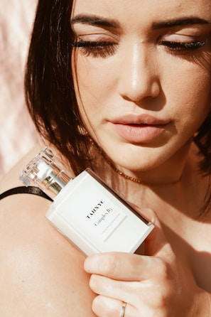 A person with closed eyes is holding a white perfume bottle close to their face, which is softly lit by natural light. The bottle is labeled 'TAHNYC Complex B3'. The person's makeup is minimal and highlights their eyelashes and lips.