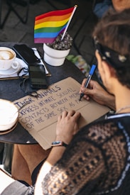 A person is writing on a cardboard sign with a blue marker. The sign contains the phrase ‘express intimate sexual contacts between men of the same sex as’. A small potted plant with a rainbow flag is placed on a table nearby along with a smartphone and a cup of coffee.