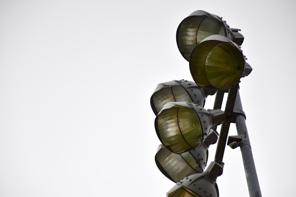 A set of large metal industrial lights mounted on a tall pole, with each light encased in a protective metal grid. The angle of the photograph creates a dramatic view of the structure against a plain, overcast sky.