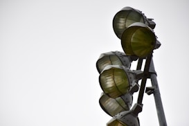 A set of large metal industrial lights mounted on a tall pole, with each light encased in a protective metal grid. The angle of the photograph creates a dramatic view of the structure against a plain, overcast sky.