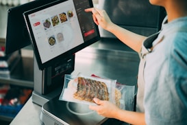 A person is interacting with a touchscreen interface on a point-of-sale terminal. The screen displays various food items with images and prices. The individual is holding a package of sliced meat while selecting options on the screen.