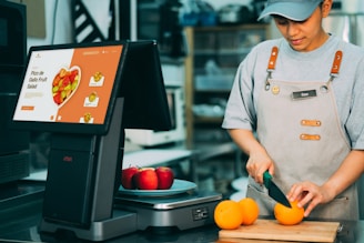 A person wearing an apron is slicing oranges on a wooden cutting board. Nearby, there is a digital screen displaying a menu for Pico de Gallo Fruit Salad with images of fruit. On the counter, there are also a few red apples.
