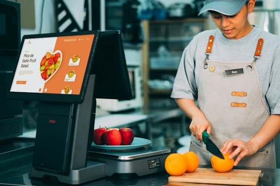 A person wearing an apron is slicing oranges on a wooden cutting board. Nearby, there is a digital screen displaying a menu for Pico de Gallo Fruit Salad with images of fruit. On the counter, there are also a few red apples.