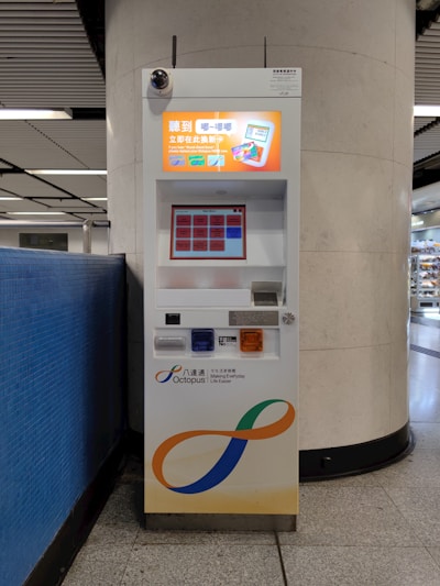 A vending machine placed between a tiled wall and a large column in a subway station. The machine has a bright display screen with text and card images, multiple buttons, and a section for card transactions. It is branded with the 'Octopus' logo and slogan 'Making Everyday Life Easier.'