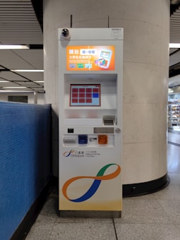 A vending machine placed between a tiled wall and a large column in a subway station. The machine has a bright display screen with text and card images, multiple buttons, and a section for card transactions. It is branded with the 'Octopus' logo and slogan 'Making Everyday Life Easier.'