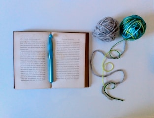 An open book lies flat with a teal crochet hook placed on it. To the right of the book, two balls of yarn, one gray and one multicolored with green and blue hues, are positioned. The yarn trails off from the balls, forming gentle curves on the white background.