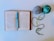 An open book lies flat with a teal crochet hook placed on it. To the right of the book, two balls of yarn, one gray and one multicolored with green and blue hues, are positioned. The yarn trails off from the balls, forming gentle curves on the white background.